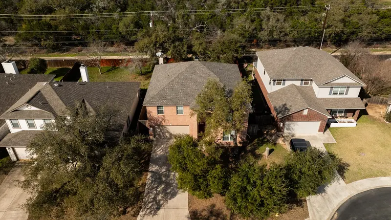 Aerial view of brick two-story home with gray asphalt shingle roof surrounded by mature trees and neighboring houses.