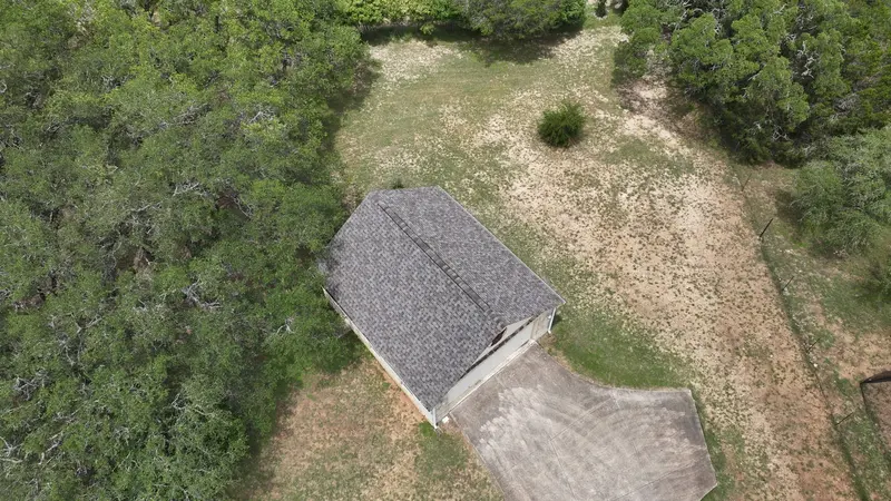 Aerial view of residential home with dark gray asphalt shingle roof surrounded by dense cedar trees and gravel clearing.