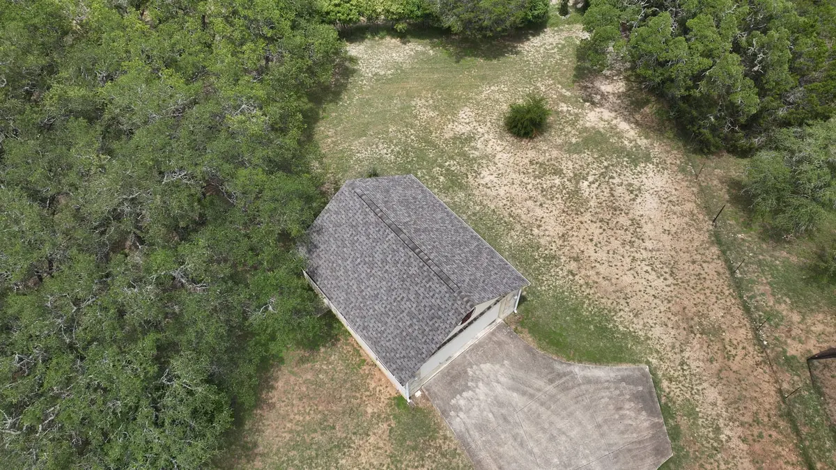 Aerial view of residential home with dark gray asphalt shingle roof surrounded by dense cedar trees and gravel clearing.