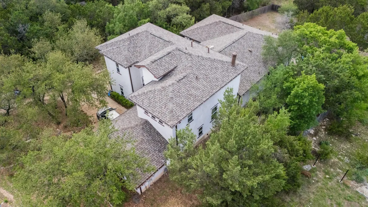 Aerial view of white modern home with gray architectural shingle roof in wooded Texas Hill Country setting