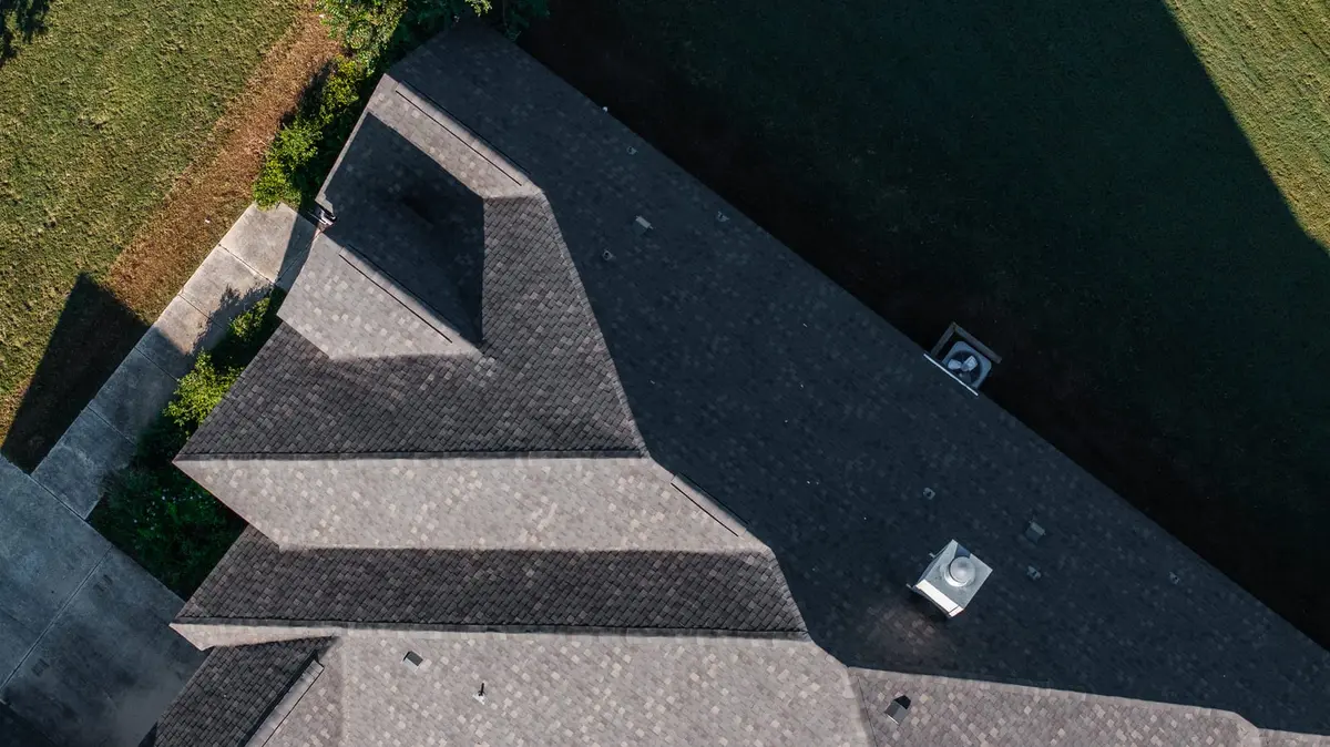 Aerial view of residential home with multi-pitched architectural shingle roof in dark gray and light tan tones, bordered by