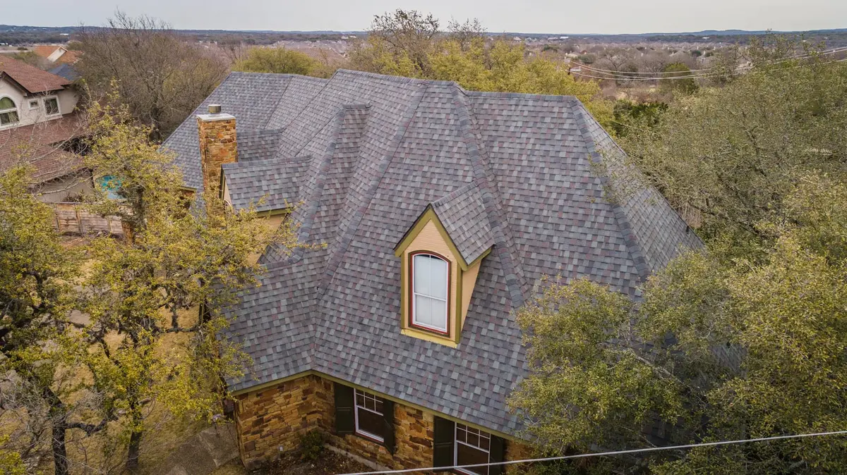 Architectural asphalt shingle roof with gray/blue tones, stone chimney, and yellow dormer window on residential home