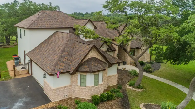 Two-story brick and stone home with brown architectural shingle roof, curved turret section, and mature oak trees