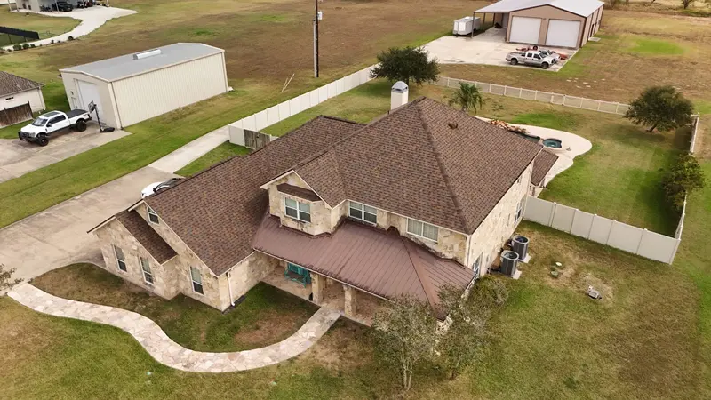 Brown architectural shingle roof on two-story stone home with metal porch covering and white vinyl fencing in rural Texas