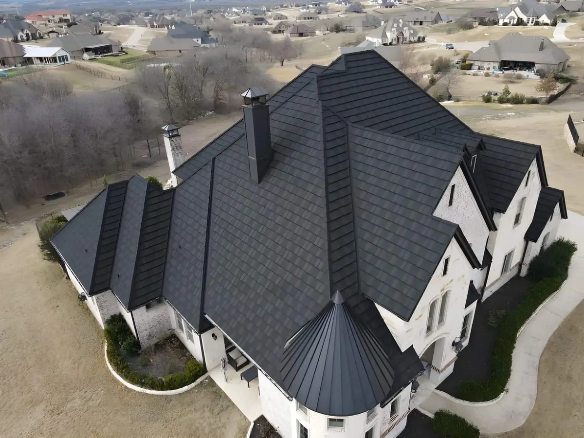 Aerial view of luxury home with black architectural shingle roof, multiple peaks, white exterior, and chimney.