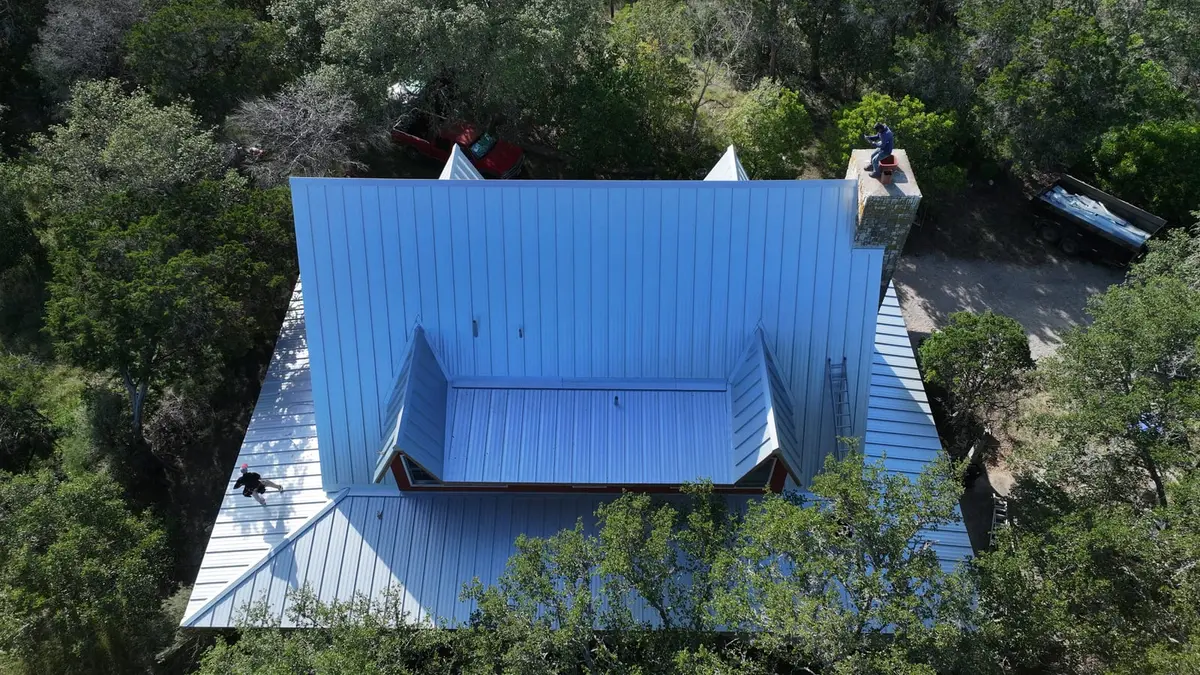 Aerial view of residential home with bright blue metal roof and white trim, surrounded by mature trees