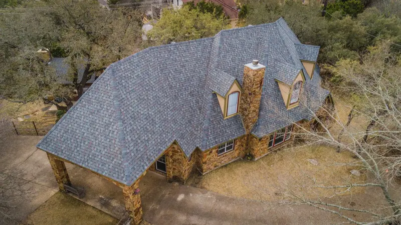 Aerial view of stone residential home with blue-gray architectural asphalt shingle roof, multiple gables, and stone
