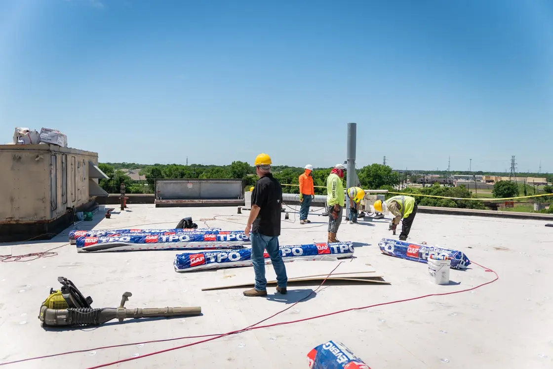 Crew staging rolls of white TPO single-ply membrane across a commercial flat roof before install