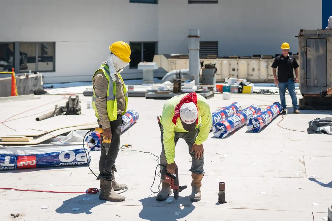 Roofing crew fastening insulation board ahead of a white TPO membrane on a flat commercial roof