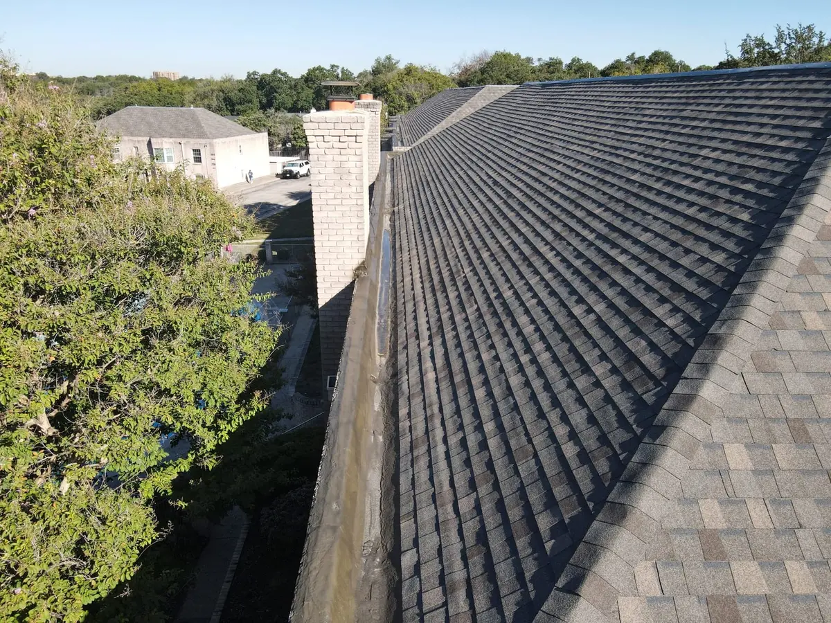 Dark architectural shingle roof with steep pitch and white brick chimney; residential structure visible in background.