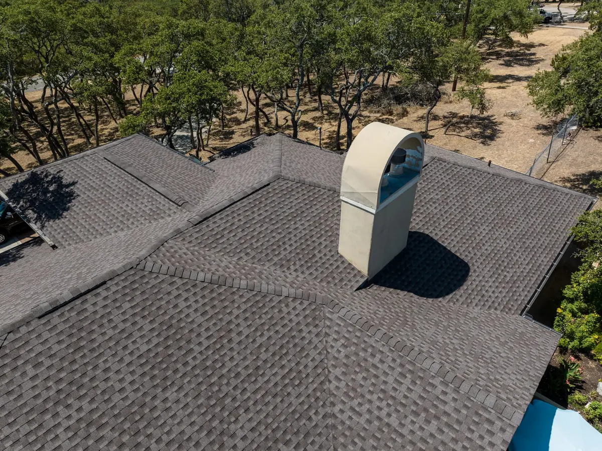 Aerial view of gray asphalt shingle roof with white cylindrical skylight, surrounded by oak trees in Texas Hill Country landscape.