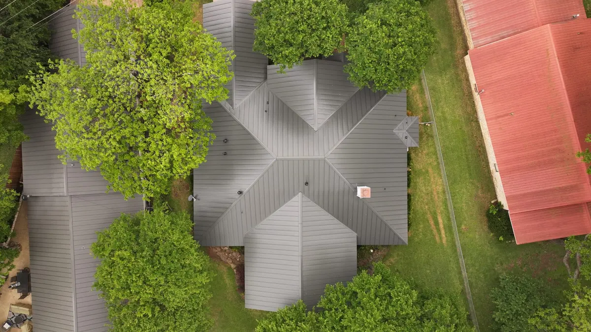 Aerial view of gray metal roof on residential home surrounded by large green trees and grass yard