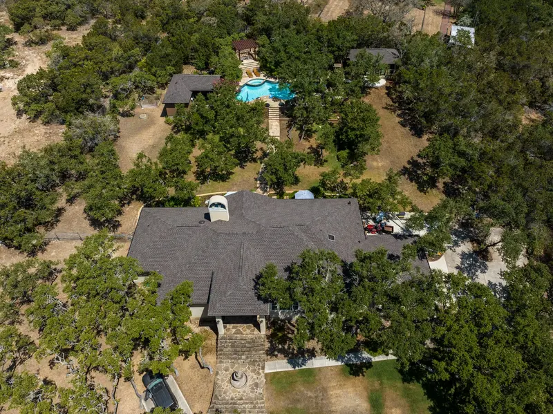 Aerial view of residential home with dark gray asphalt shingle roof surrounded by oak trees in Texas Hill Country setting