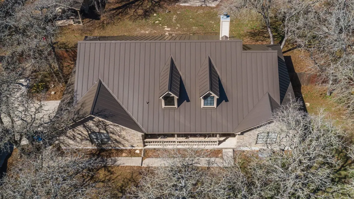Aerial view of residential home with gray metal standing-seam roof, two dormer windows, stone foundation, surrounded by