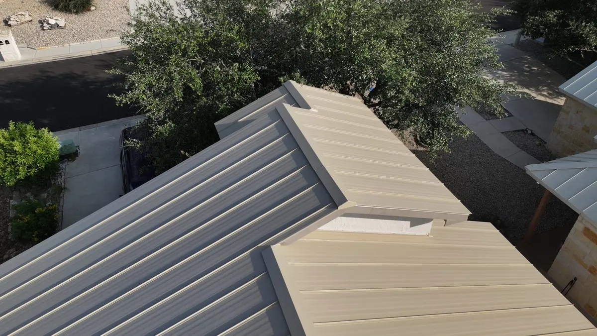 Aerial view of metal standing-seam residential roof with tan/beige panels and gray trim, surrounded by green trees and