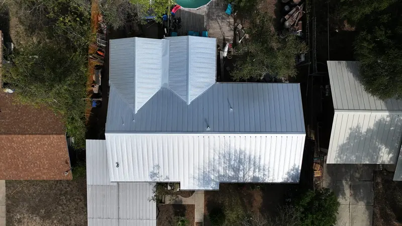 Aerial view of residential house with new metal standing-seam roof installation, white and gray panels, surrounding mature
