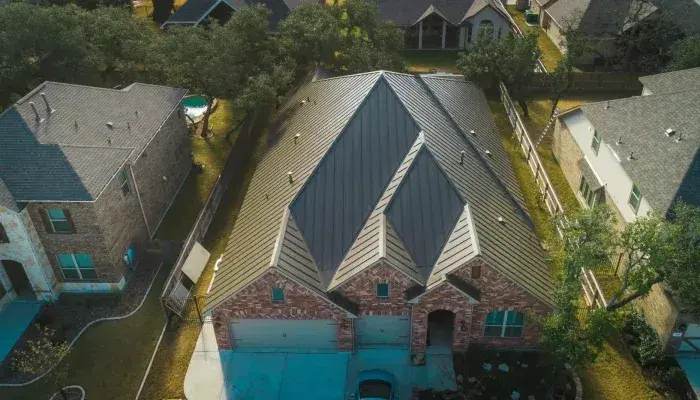 Aerial view of brick home with dark metal standing seam roof featuring distinctive peaked gables and turquoise garage doors