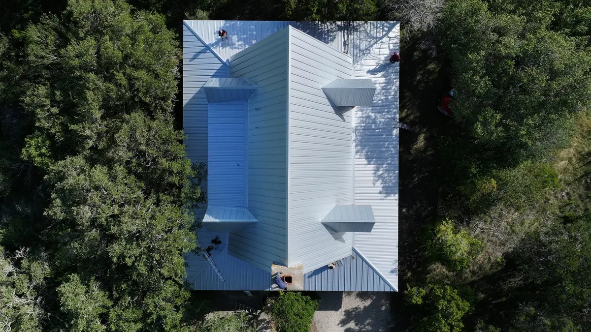 Aerial view of residential metal roof installation with standing seam panels, blue and white finishes, surrounded by dense