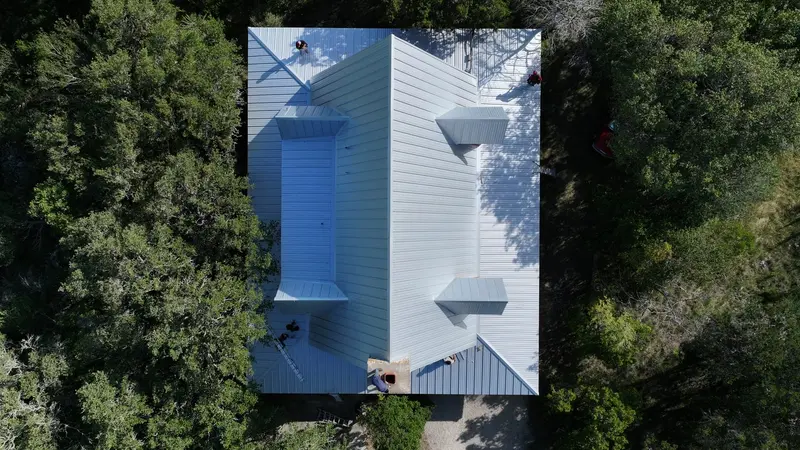 Aerial view of residential metal roof installation with standing seam panels, blue and white finishes, surrounded by dense