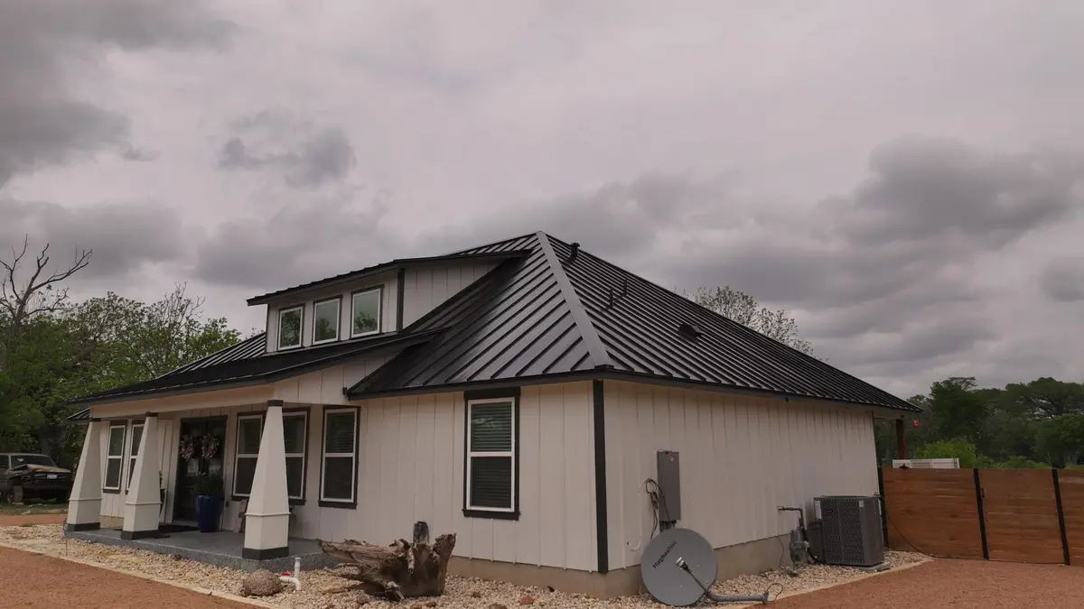 Modern residential home with black metal standing-seam roof and steep peak, cream vertical siding, multiple windows.
