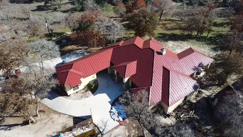 Aerial view of residential home with red metal standing-seam roof, complex multi-peak design, surrounded by trees on wooded