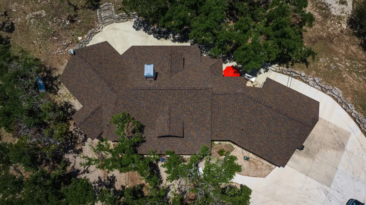 Aerial view of residential home with brown architectural shingle roof, white trim, concrete driveway, and surrounding oak