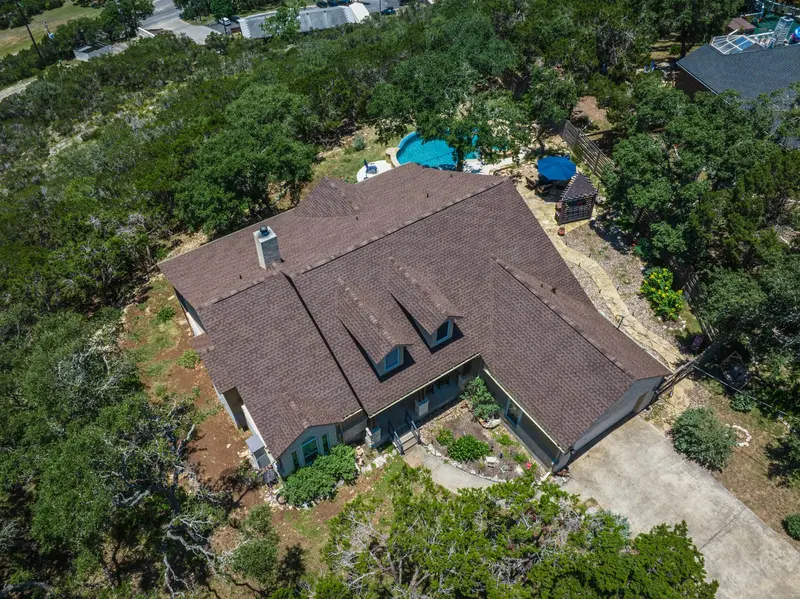 Aerial view of residential home with dark brown asphalt shingle roof, two roof vents, surrounded by oak trees and blue pool