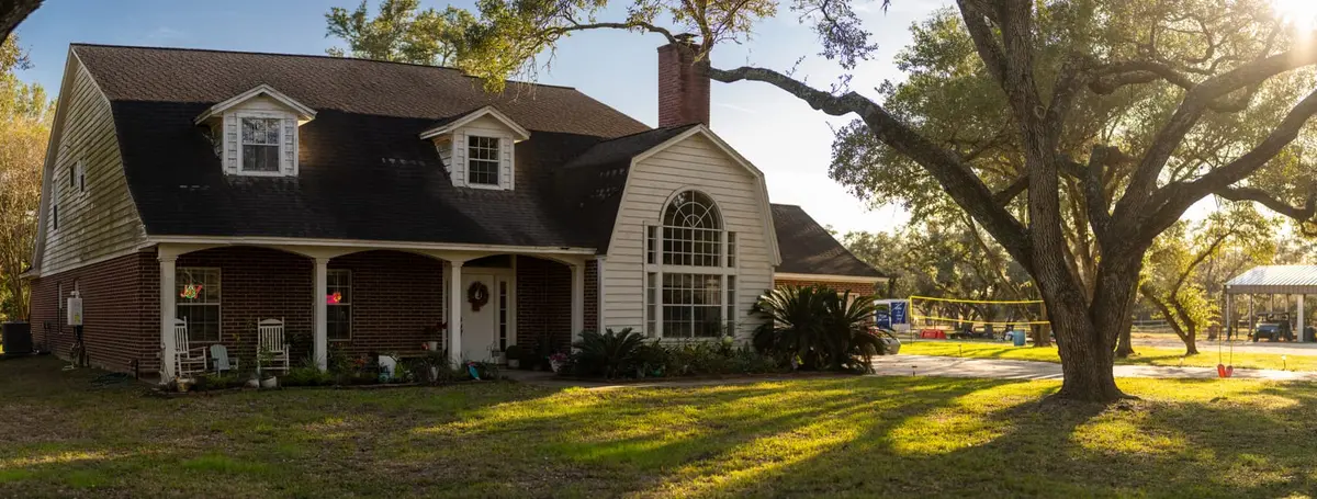 Two-story brick and vinyl residential home with dark asphalt shingle roof, dormers, and large oak tree in front yard