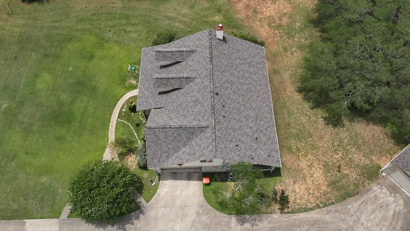 Aerial view of residential house with gray architectural shingle roof, two black horizontal vents, chimney, curved