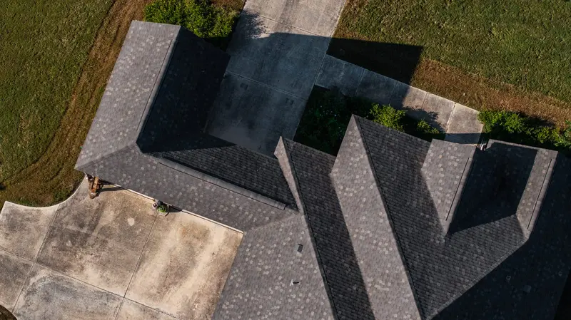 Aerial view of residential home with multiple peaked roofs covered in dark gray architectural shingles and strong shadow