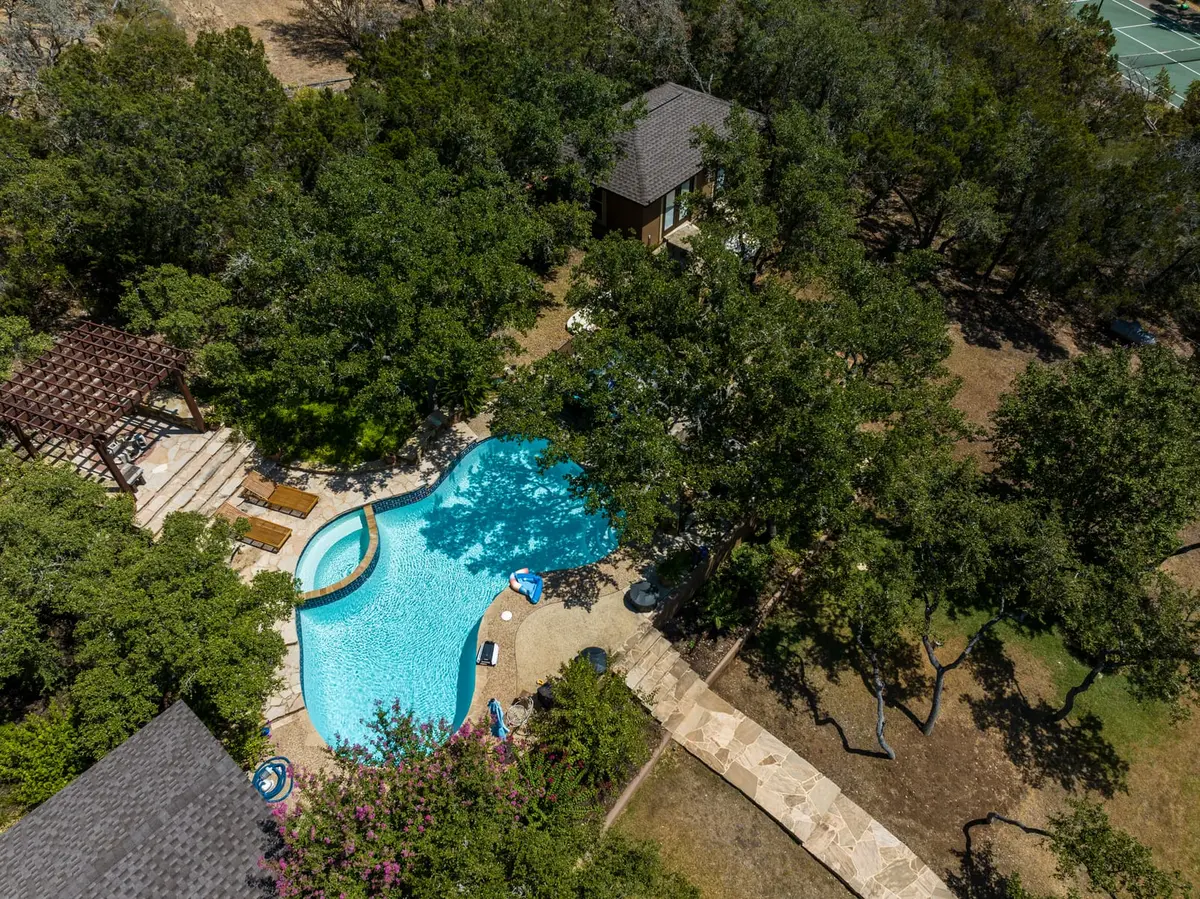 Aerial view of residential home with dark gray tile roof nestled among mature oak trees with curved swimming pool and patio