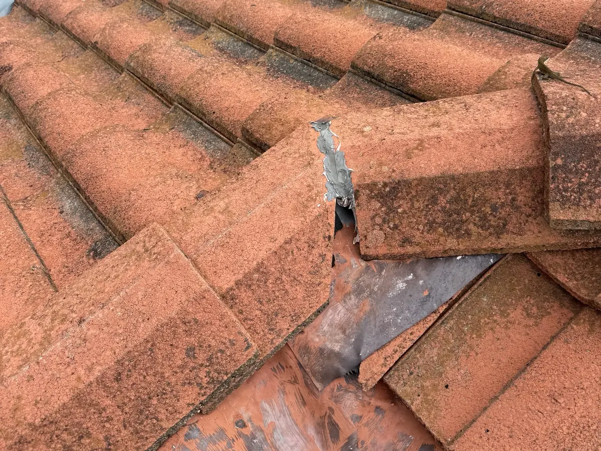 A broken roof tile in a valley patched with gray sealant, with tiles shifted off the metal valley flashing underneath