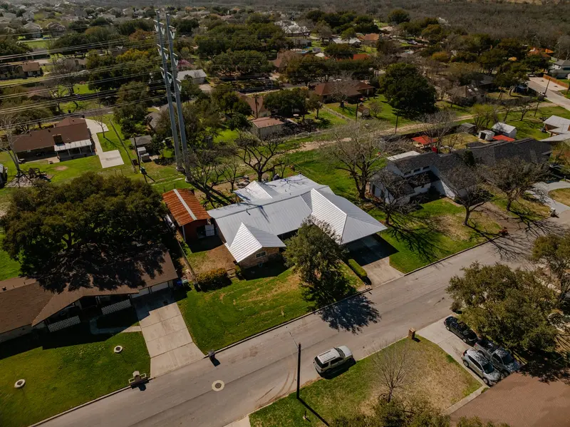 Aerial view of residential home with white metal standing-seam roof and brick exterior on tree-lined suburban lot